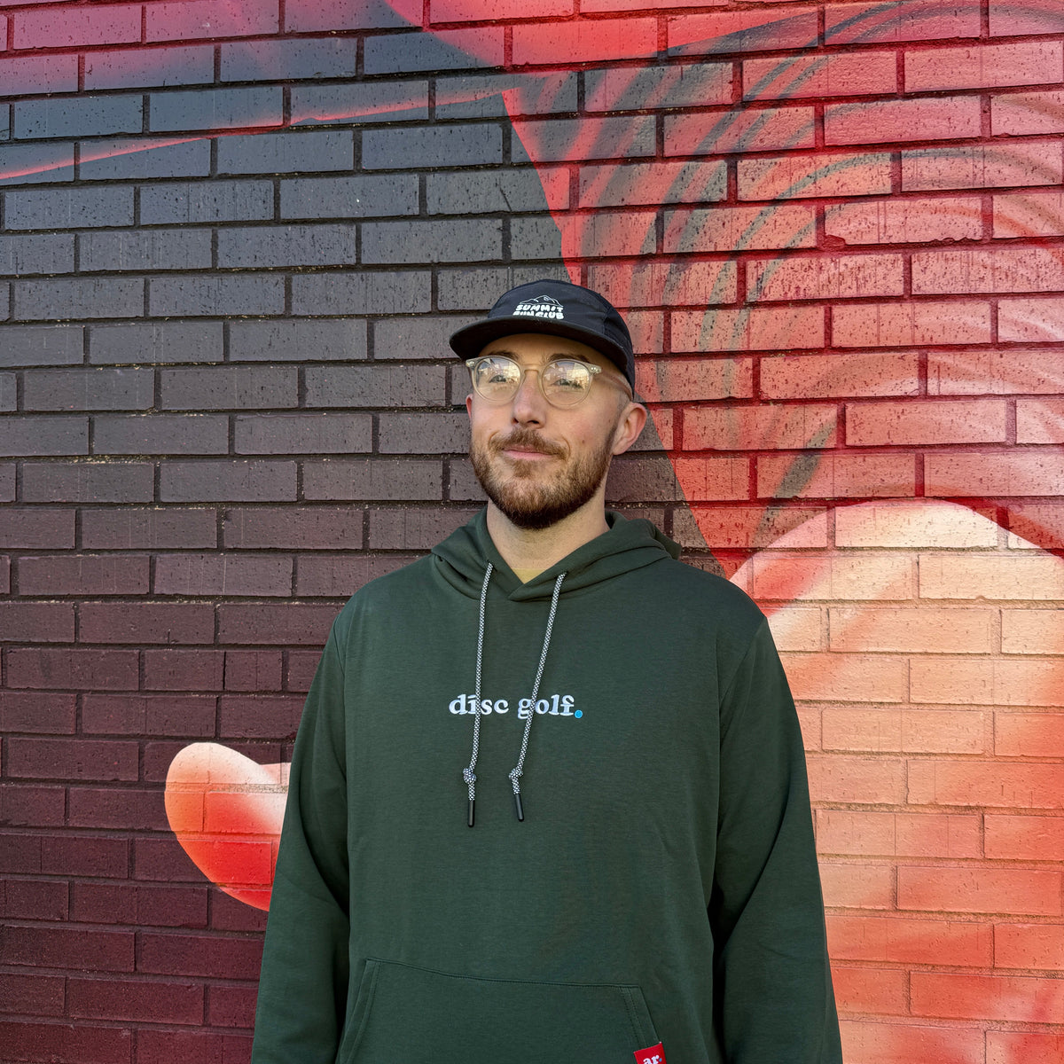 A man wearing a dark green 'disc golf.' hoodie, a black cap, and glasses stands in front of a brick wall with a red and black graffiti mural.