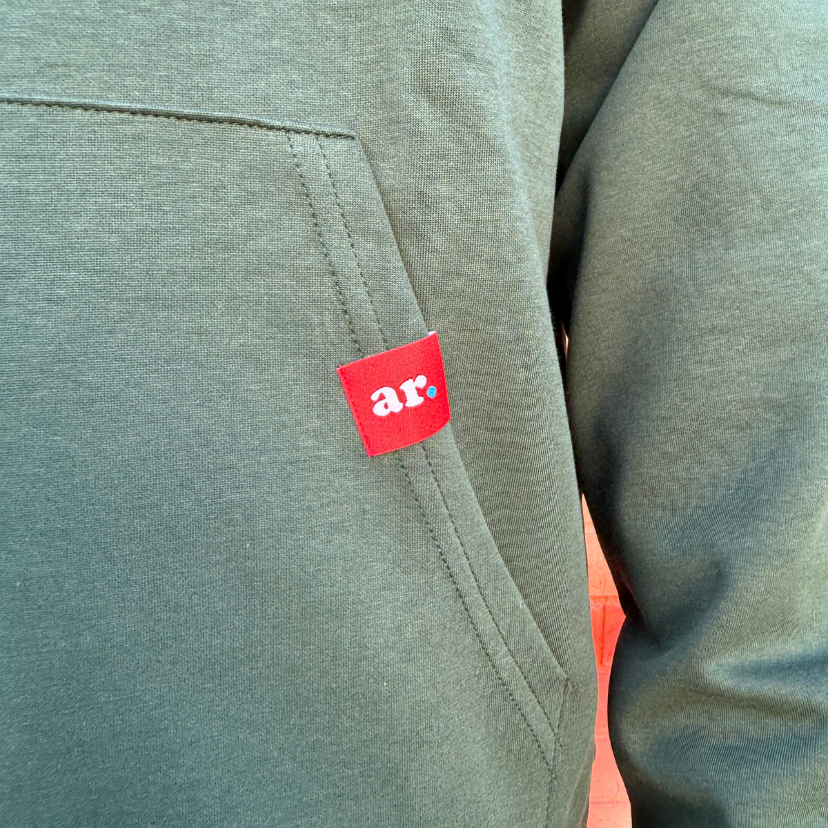 Close-up of a heathered sage green hoodie with a red brand patch featuring the white text 'ar.' on the chest pocket.