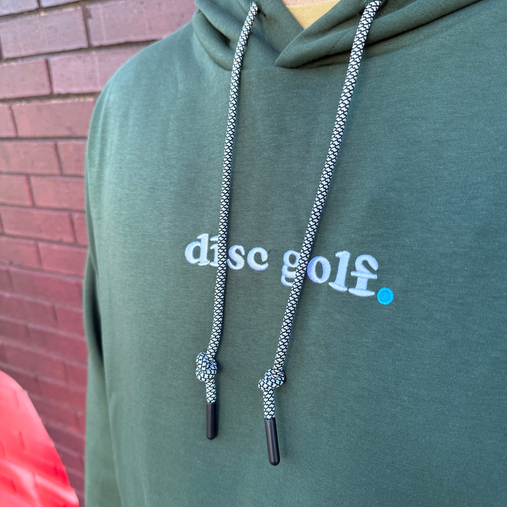 A close-up of a person wearing a dark green hoodie with 'disc golf.' printed in white across the chest, standing in front of a red brick wall.