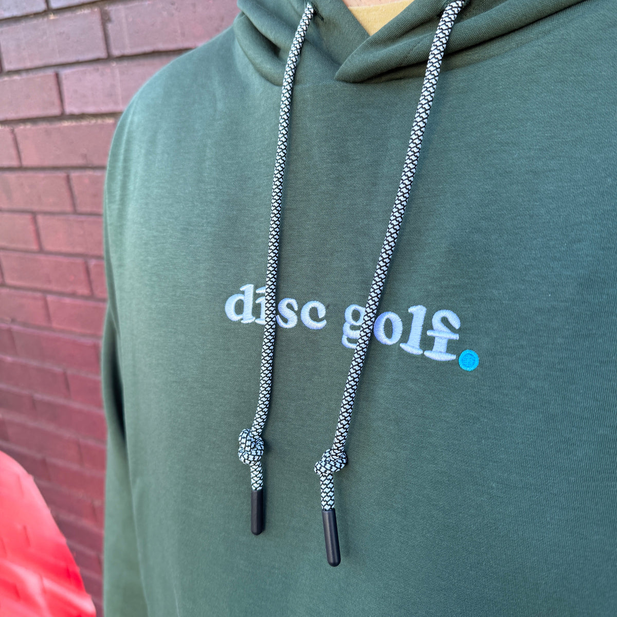 A close-up of a person wearing a dark green hoodie with 'disc golf.' printed in white across the chest, standing in front of a red brick wall.