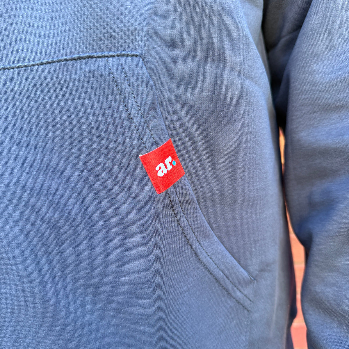 Close-up of a heathered slate blue hoodie featuring a small red square patch with the white lowercase letters 'ar.' near the pocket.