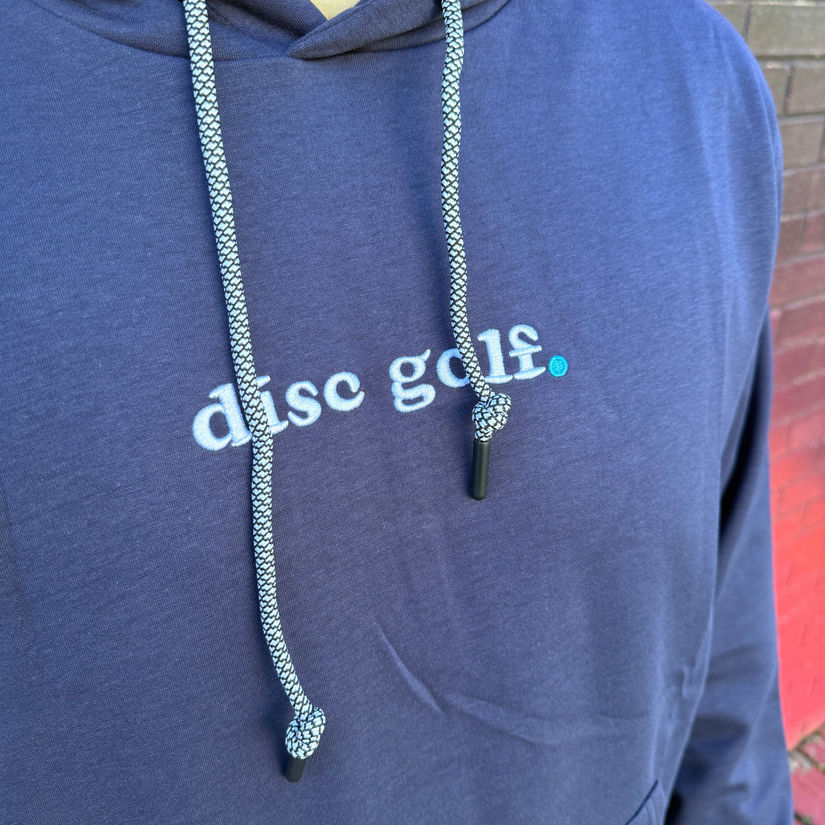 A close-up of a person wearing a navy blue hoodie with 'disc golf.' printed across the chest in white and a small teal dot.